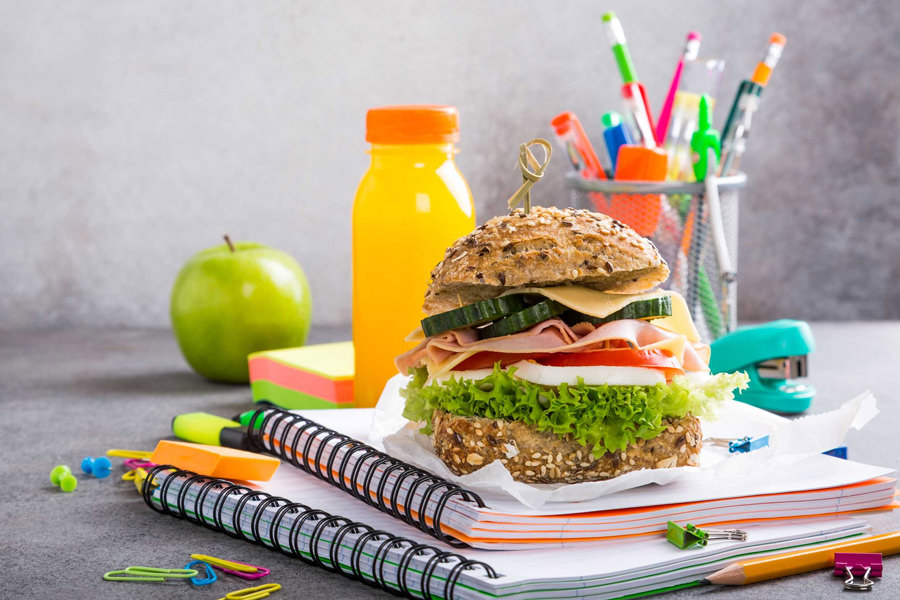 Belegtes Brötchen liegt auf Schulheften neben Getränk und Schulsachen